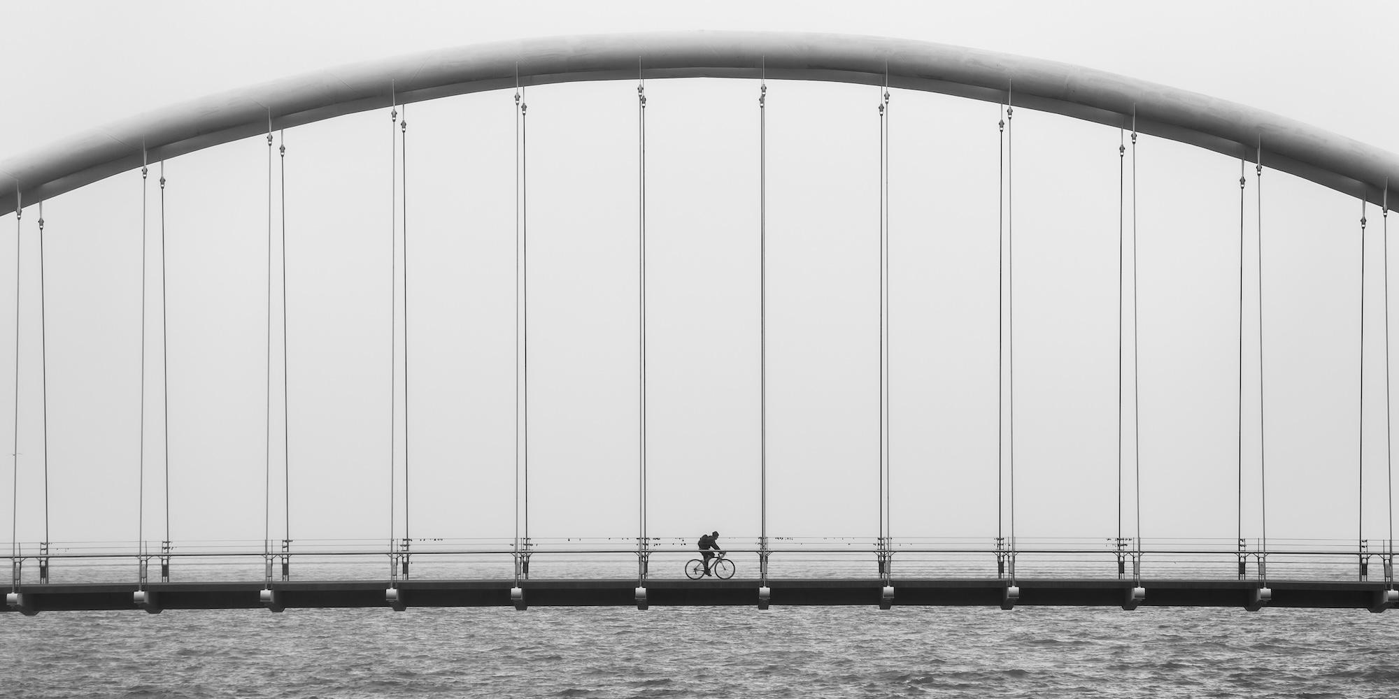 Cover image for this post: Grayscale photo of a person biking over a bridge, over water.